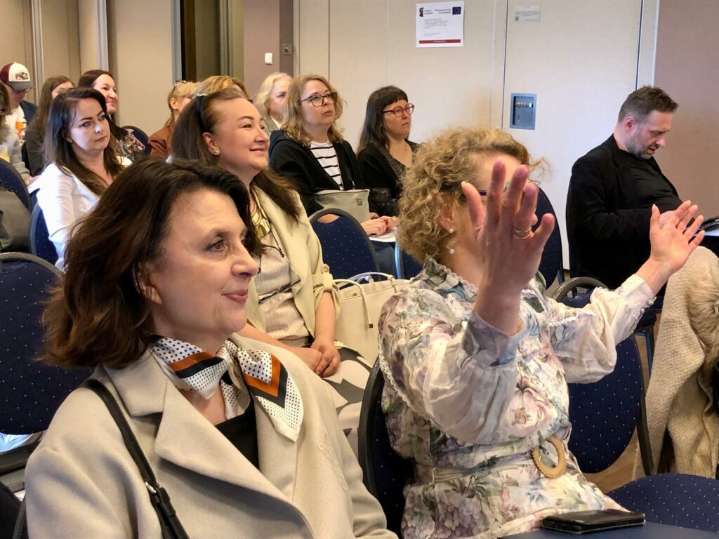 Sala konferencyjna. Uczestnicy konferencji słuchają prezentacji. Uśmiechają się. Anna Janus podnosi ręce,