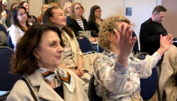 Sala konferencyjna. Uczestnicy konferencji słuchają prezentacji. Uśmiechają się. Anna Janus podnosi ręce.