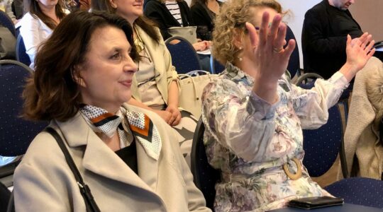 Sala konferencyjna. Uczestnicy konferencji słuchają prezentacji. Uśmiechają się. Anna Janus podnosi ręce.