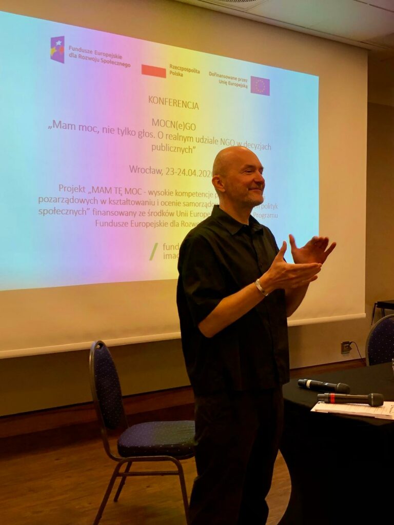 Sala konferencyjna. Na tle prezentacji poświęconej projektowi “Mam Tę Moc” stoi Piotr Kuźniak z Fundacji Imago. Uśmiecha się, klaszcze.