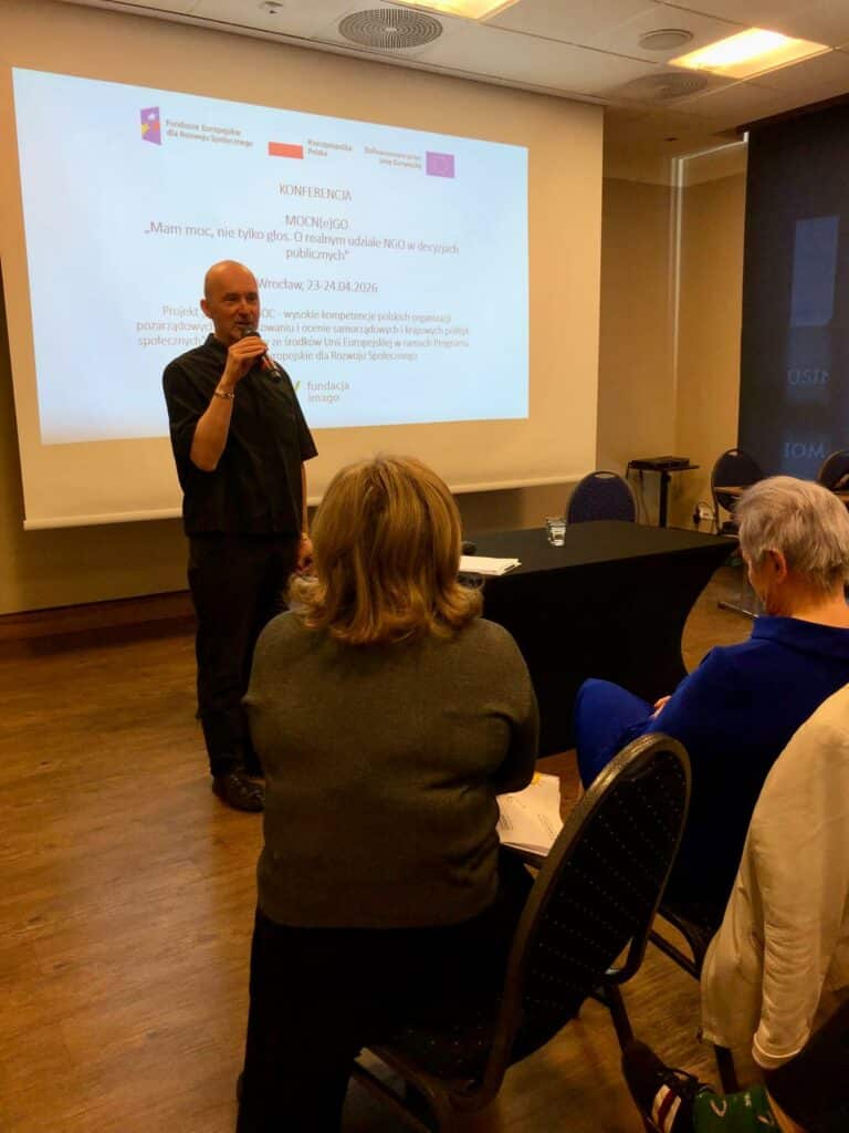 Sala konferencyjna. Na tle prezentacji poświęconej projektowi “Mam Tę Moc” stoi Piotr Kuźniak z Fundacji Imago. W ręce ma mikrofon. Przed nim uczestnicy konferencji.