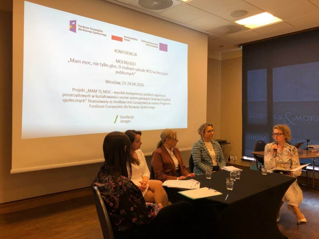 Pięć uczestniczek i prowadzących panelu dyskusyjnego z uczestniczkami projektu “Mam Tę Moc” w sali konferencyjnej, przy stoliku. Anna Janus z Fundacji Imago trzyma mikrofon, rozpoczynając dyskusję. W tle ekran z prezentacją.