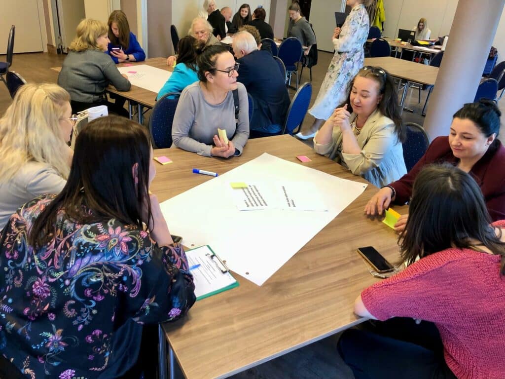 Zdjęcie w sali konferencyjnej. Przy stolikach siedzą grupy uczestników konferencji z moderatorami z Fundacji Imago. Wykonują zadania - przed nimi leżą duże, białe kartki i małe karteczki.