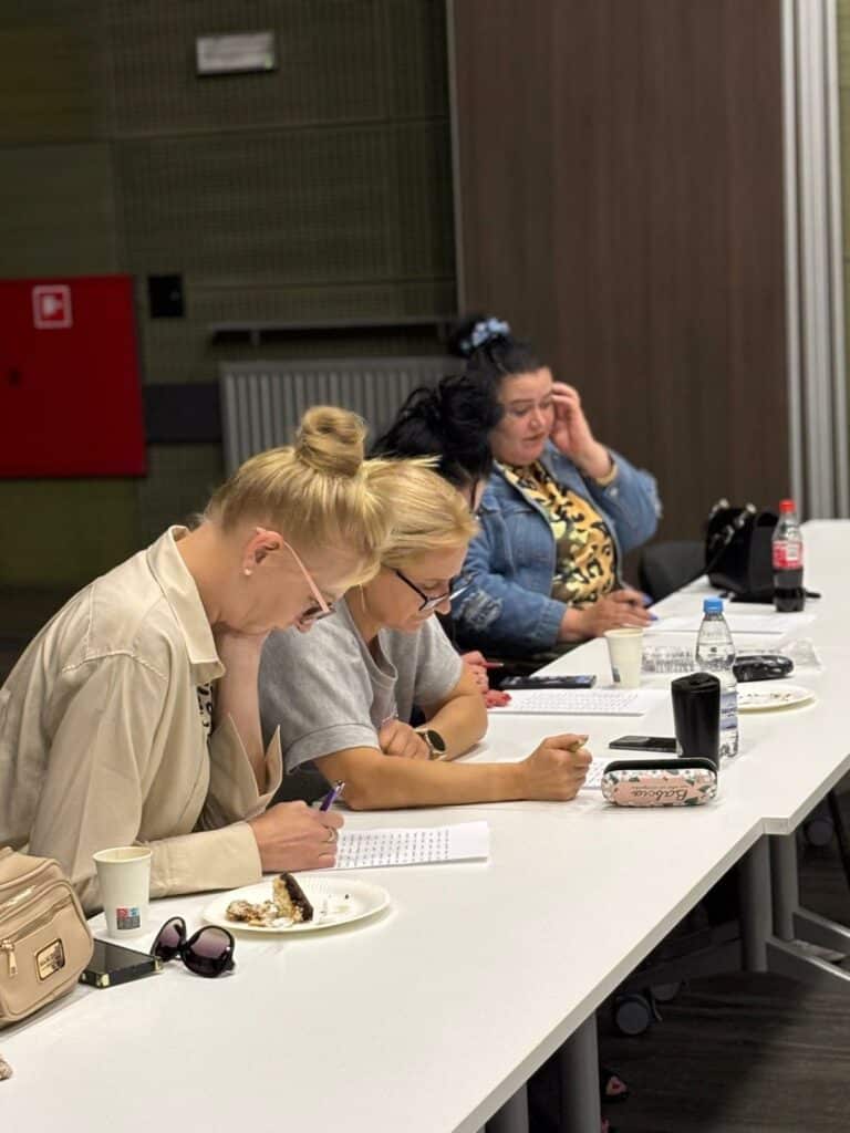 Sala szkoleniowa, cztery uczestniczki siedzą przy stole, przed nimi kartki papieru, kobiety wypełniają zadanie szkoleniowe. 
