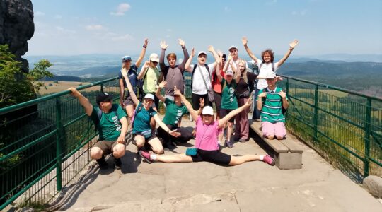 14-osobowa grupa pozuje do zdjęcia na szczycie góry, unosząc ręce w górę.