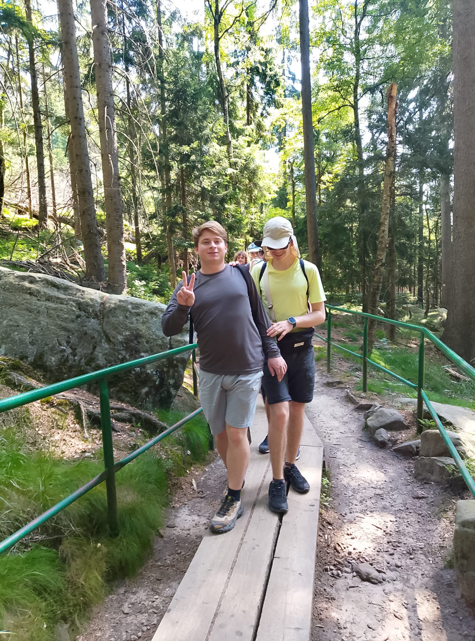 Dwie osoby na drewnianej ścieżce w lesie. Chłopak unosi rękę w górę w geście zwycięstwa. Jego ręki trzyma się uśmiechnięta dziewczyna, która prawdopodobnie ma niepełnosprawność wzroku.