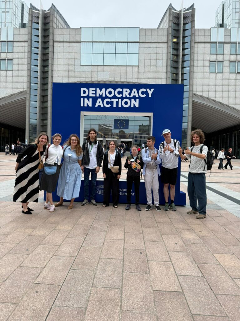 9-osobowa grupa na tle budynku Parlamentu Europejskiego w Brukseli. To uczestnicy, wolontariusze i koordynatorzy projektu "Points of konekszyn". Za nimi duża rama do zdjeć z napisem "Demokrasi in ekszyn".