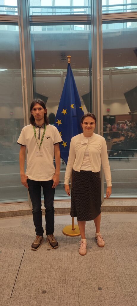 Uczestnik i uczestniczka projektu "Points of konekszyn" pozują do zdjęcia w budynku Parlamentu Europejskiego w Brukseli. Za nimi na stojaku duża flaga Unii Europejskiej.