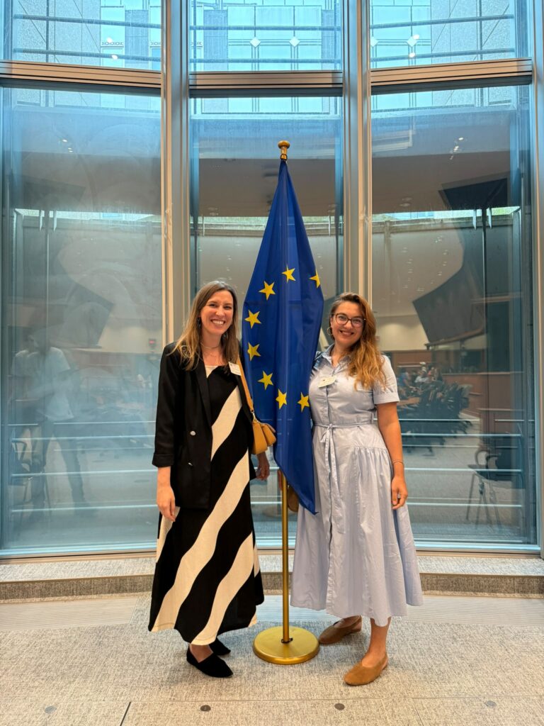 Martyna Rangno i Anna Dadura z Fundacji Imago pozują do zdjęcia w budynku Parlamentu Europejskiego w Brukseli. Za nimi na stojaku duża flaga Unii Europejskiej.