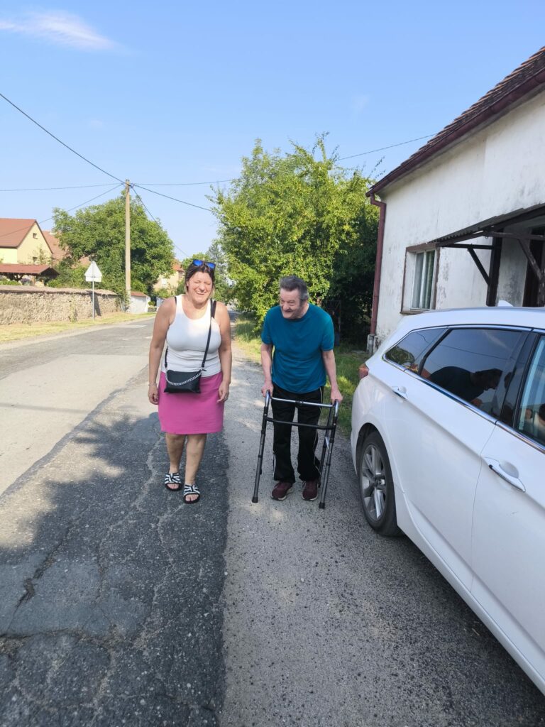Na słonecznej ulicy przed białym budynkiem stoją kobieta i mężczyzna. Kobieta uśmiecha się do aparatu; ma na sobie białą bluzkę bez rękawów, różową spódnicę i małą czarną torebkę. Mężczyzna stoi obok, opierając się na chodziku, i również się uśmiecha. Po prawej stronie znajduje się biały samochód. W tle widać drzewa, domy, drogę oraz bezchmurne, błękitne niebo.