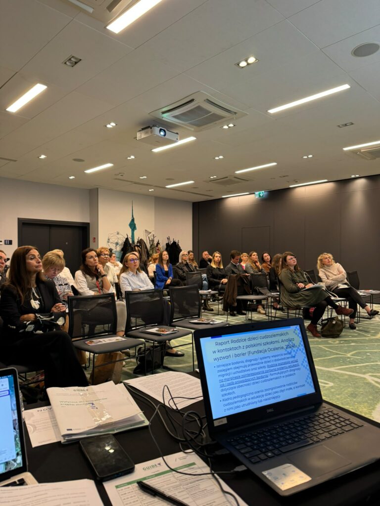 Sala konferencyjna. Na krzesłach siedzi ponad 20 uczestników konferencji Gajd. Słuchają prezentacji. Jeden ze slajdów widać na ustawionym przed nimi na stoliku komputerze, z którego puszczana jest prezentacja.