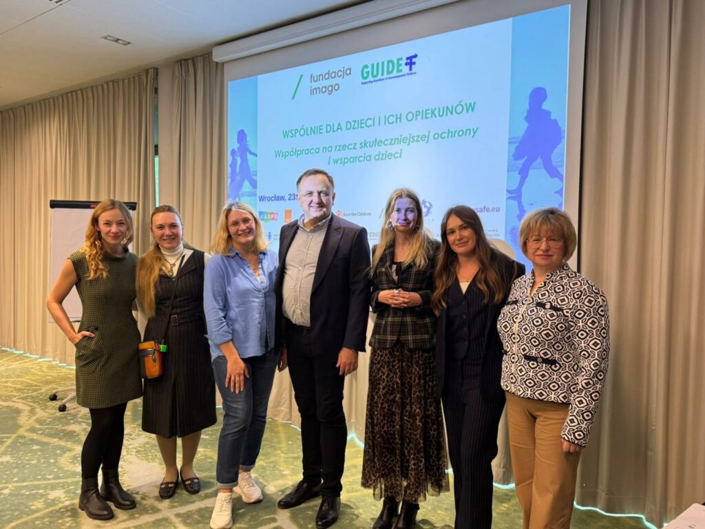 7 osób pozuje do zdjęcia w sali konferencyjnej na tle dużego ekranu z logotypami projektu Gajd oraz tytułem konferencji: "Wspólnie dla dzieci i ich opiekunów. Współpraca na rzecz skuteczniejszej ochrony i wsparcia dzieci". Wśród nich są Marija Pavlova i Magda Mielczarek z Fundacji Imago.