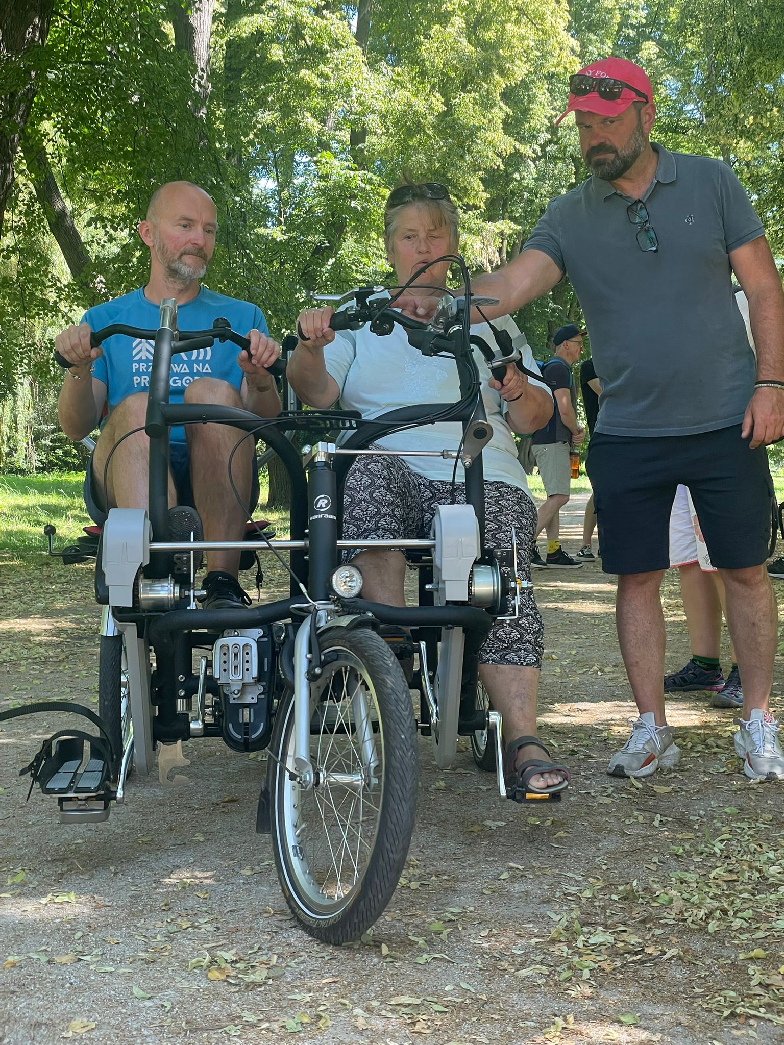 Zdjęcie trzech osób w parku. Dwie z nich siedzą obok siebie na specjalnym rowerze - tandemie. To Piotr Kuźniak, prezes Fundacji Imago oraz Gabriela Trela-Kiljan, osoba z niepełnosprawnością. Obok stoi mężczyzna, który wyjaśnia coś na temat roweru.