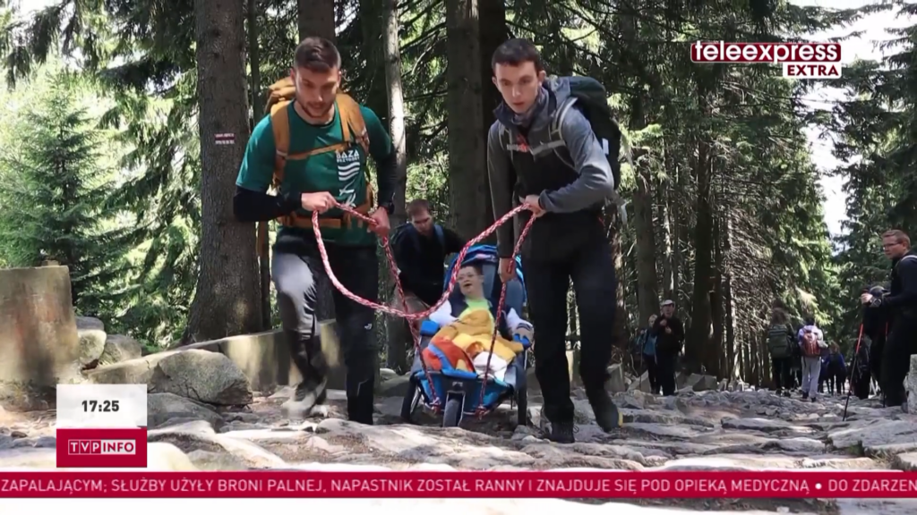 Stopklatka z programu Teleexpress Extra. Na zdjęciu droga pod górę w lesie. Dwóch młodych mężczyzn ciągnie wózek trekkingowy, w którym jest chłopak z niepełnosprawnością. W tle inni uczestnicy wyprawy.