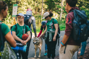 Na zdjęciu grupa osób na wyprawie w górach. Każdy ma plecak i potrzebne rzeczy. Jedna dziewczyna głaszcze psa.