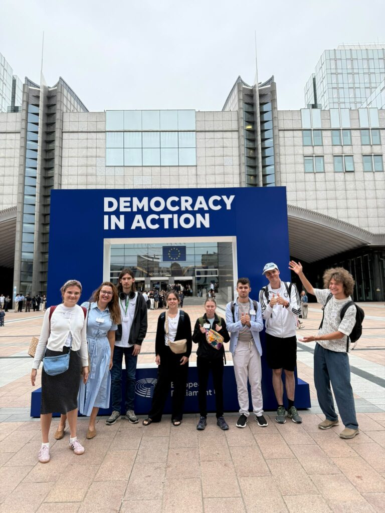 8-osobowa grupa na tle budynku Parlamentu Europejskiego w Brukseli. To uczestnicy, wolontariusze i koordynatorzy projektu "Poins of konekszyn". Zanimi duża rama do zdjeć z napisem "Demokrasi in ekszyn".