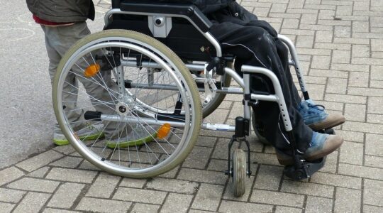 Osoba na wózku dla osób z niepełnosprawnością i człowiek, który prowadzi wózek. Zdjęcie na świeżym powietrzu. Nie widać twarzy fotografowanych.