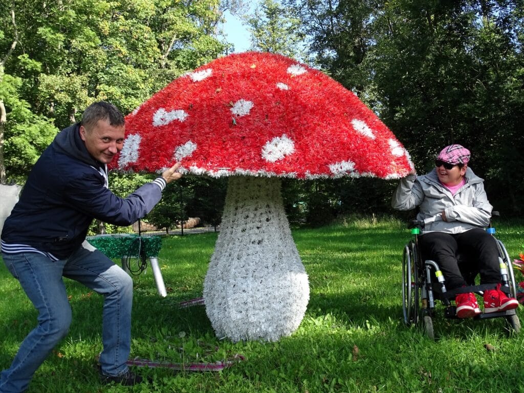 Dwie osoby pozują do zdjęcia obok dużej figury grzyba - muchomora. Jedna z nich jest na wózku dla osoby z niepełnosprawnością. Uśmiechają się.