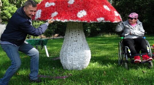 Dwie osoby pozują do zdjęcia obok dużej figury grzyba - muchomora. Jedna z nich jest na wózku dla osoby z niepełnosprawnością. Uśmiechają się.