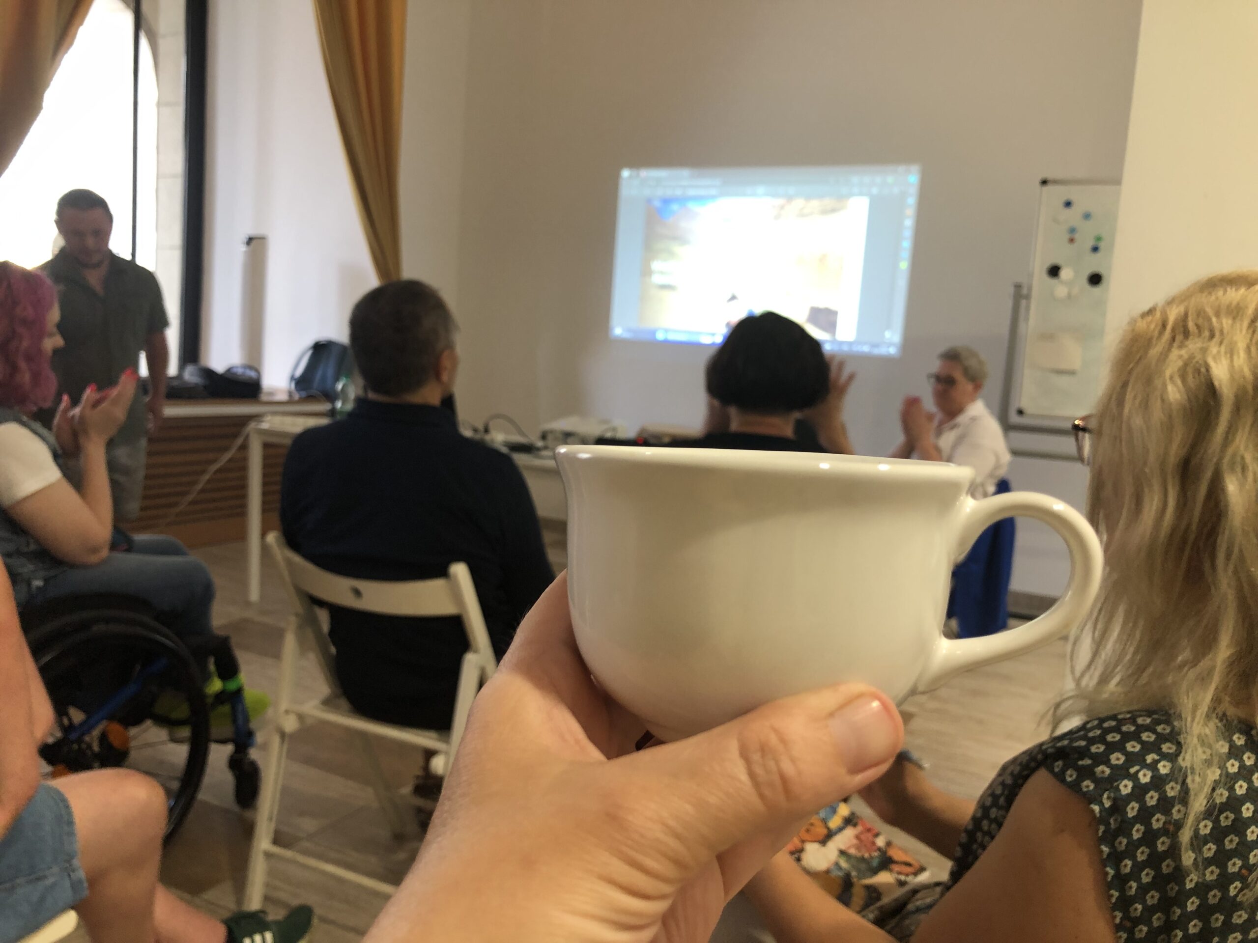 Zdjęcie wykonane w czasie spotkania Cafe EPD. Na pierwszym planie ręka trzymająca filiżankę, w tle uczestnicy spotkania i prelegenci.