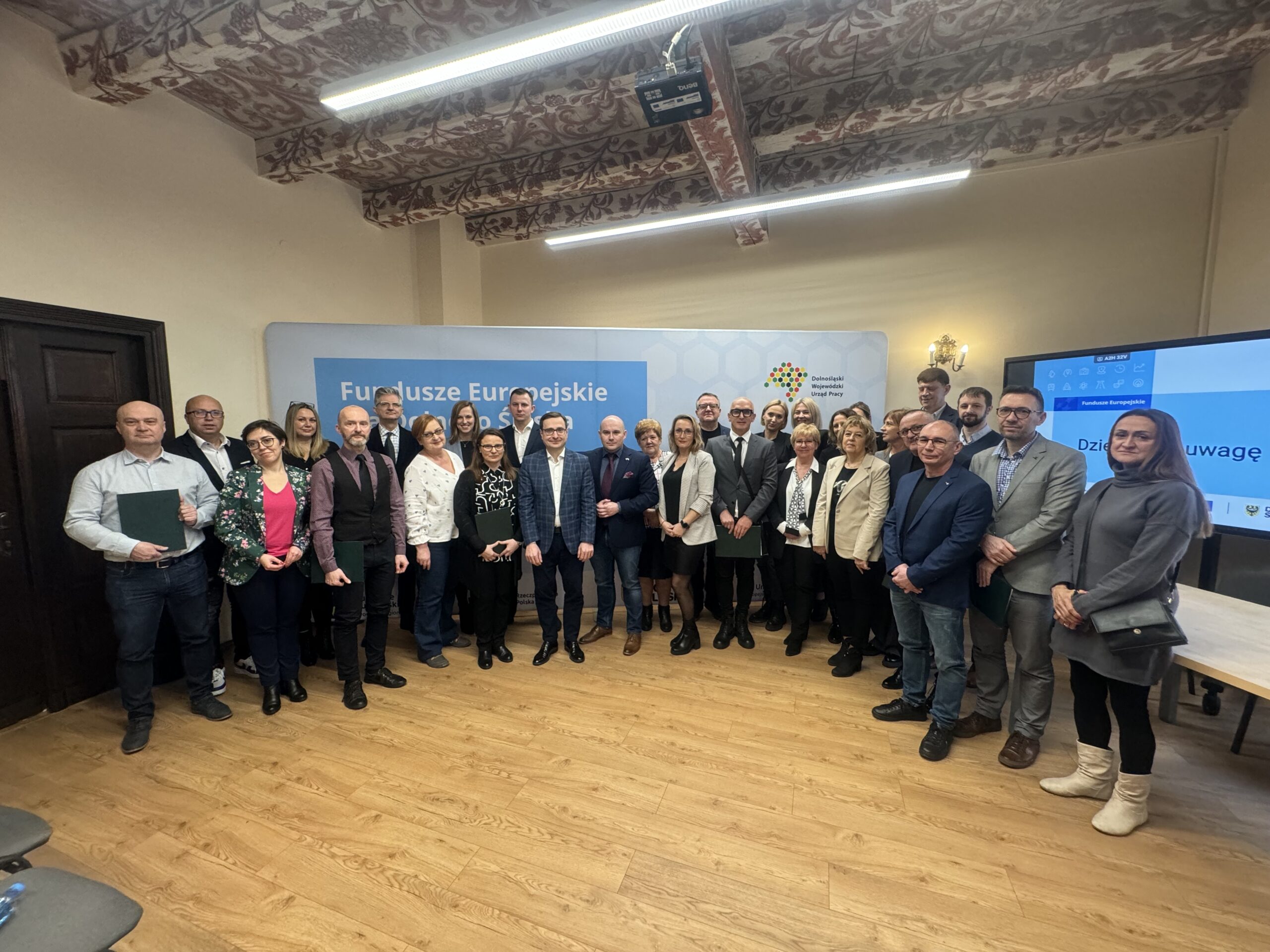 Fotografia wykonana w sali, gdzie odbywała się uroczystość podpisania umów na dofinansowanie projektów. Na środku sali, na tle “ścianki” z napisem “Fundusze Europejskie” i z logotypami stoją Bartosz Kotecki, dyrektor Dolnośląskiego Wojewódzkiego Urzędu Pracy oraz Marcin Krzyżanowski, Wicemarszałek Województwa Dolnośląskiego, obok nich beneficjenci, którzy właśnie podpisali umowy na dofinansowanie realizacji projektów, wśród nich jest Piotr Kuźniak z Fundacji Imago. Do zdjęcia pozuje około 30 osób.Salę, gdzie odbywa się spotkanie, wyróżnia ozdobny sufit, zdobiony ornamentem z motywem kwiatów, w kolorze bordowym na białym tle.