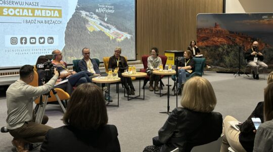 Zdjęcie zrobione w czasie Dolnośląskiego Kongresu Dostępności, w czasie panelu "Turystyka Włączająca a Jakość Życia". Przy stolikach siedzi 6 panelistów, w tle prowadzące kongres oraz tłumacz języka migowego. Za nimi duże zdjęcie atrakcji turystycznej Dolnego Śląska.