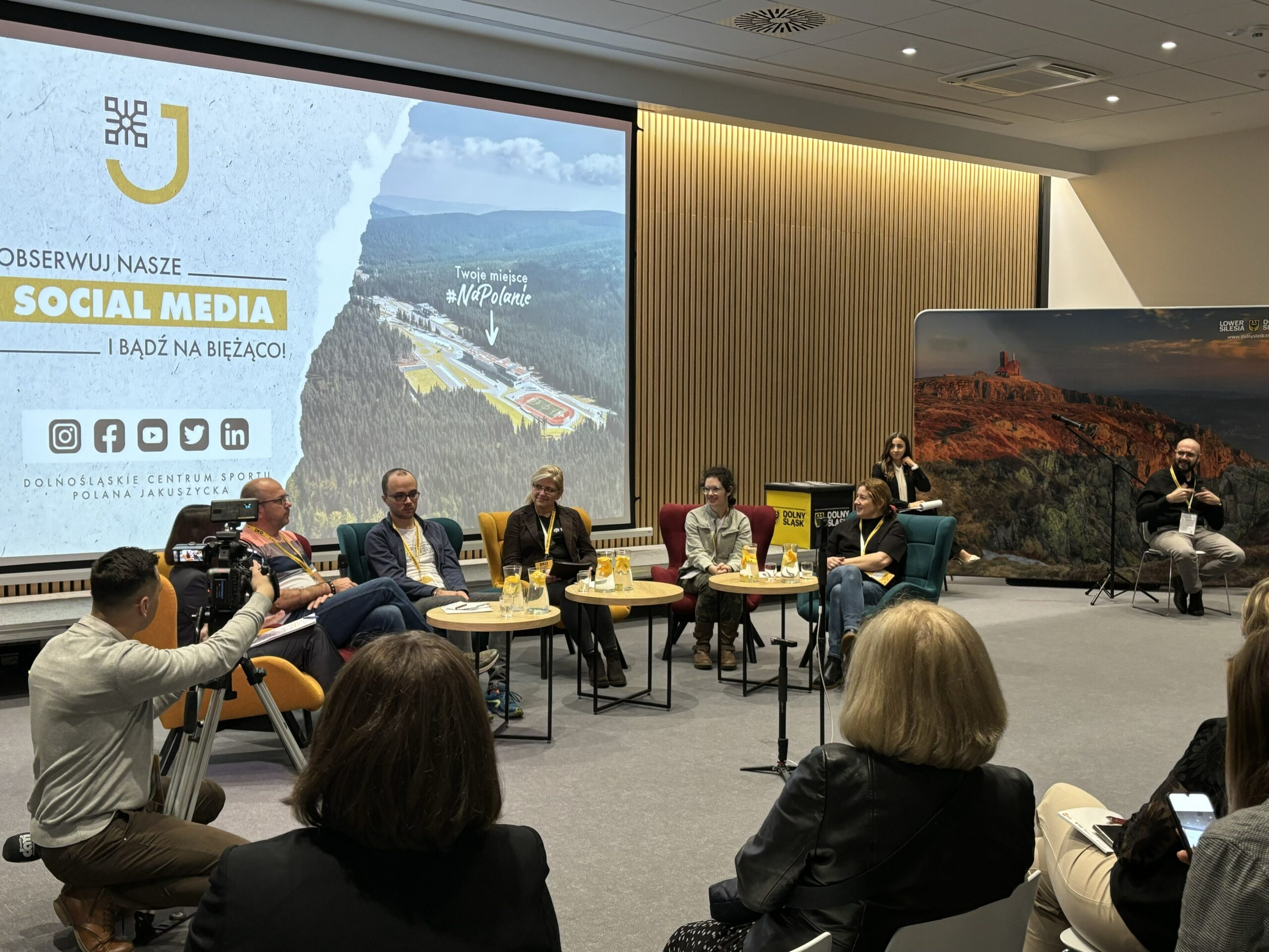 Zdjęcie zrobione w czasie Dolnośląskiego Kongresu Dostępności, w czasie panelu "Turystyka Włączająca a Jakość Życia". Przy stolikach siedzi 6 panelistów, w tle prowadzące kongres oraz tłumacz języka migowego. Za nimi duże zdjęcie atrakcji turystycznej Dolnego Śląska.