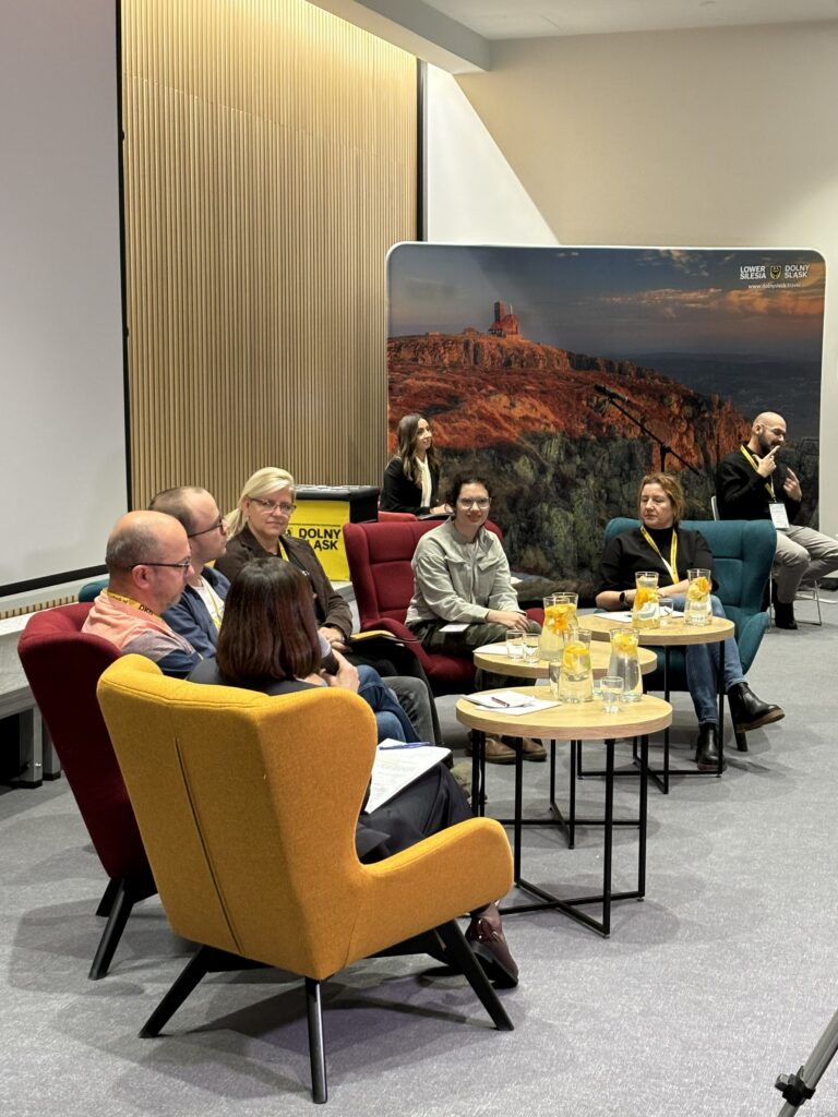 Zdjęcie zrobione w czasie Dolnośląskiego Kongresu Dostępności, w czasie panelu "Turystyka Włączająca a Jakość Życia". Przy stolikach siedzi 6 panelistów, w tle prowadzące kongres oraz tłumacz języka migowego. Za nimi duże zdjęcie atrakcji turystycznej Dolnego Śląska. 