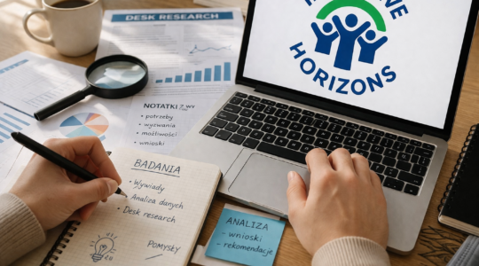 Biurko, na biurku notatki i ekran komputera z wyświetlonym logotypem projektu Inklusif Horizons. Na zdjęciu widać też ręce osoby siedzącej przy komputerze.