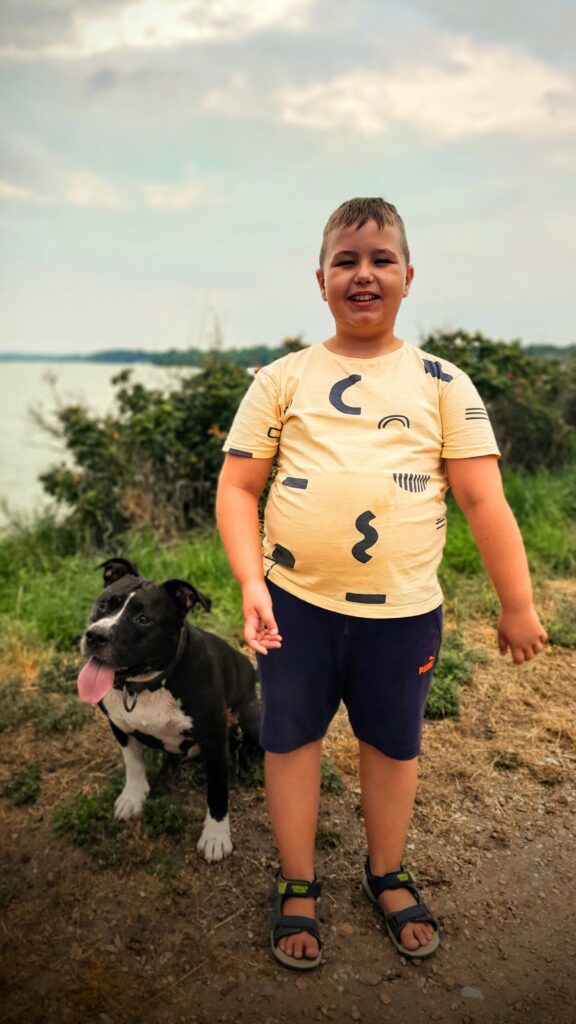 Uśmiechnięty chłopiec, obok niego pies. Zdjęcie na tle zieleni i rzeki lub jeziora. Chłopiec ma na sobie koszulkę na krótki rękaw i szorty.