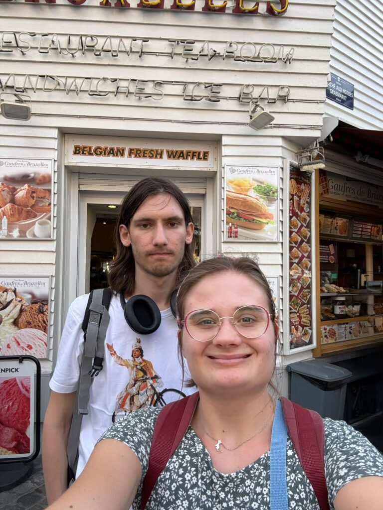 Dwie młode osoby - kobieta i mężczyzna - pozują do zdjęcia w Brukseli, na tle budki sprzedającej wafle belgijskie. To uczestnicy projektu "Poins of konekszyn".