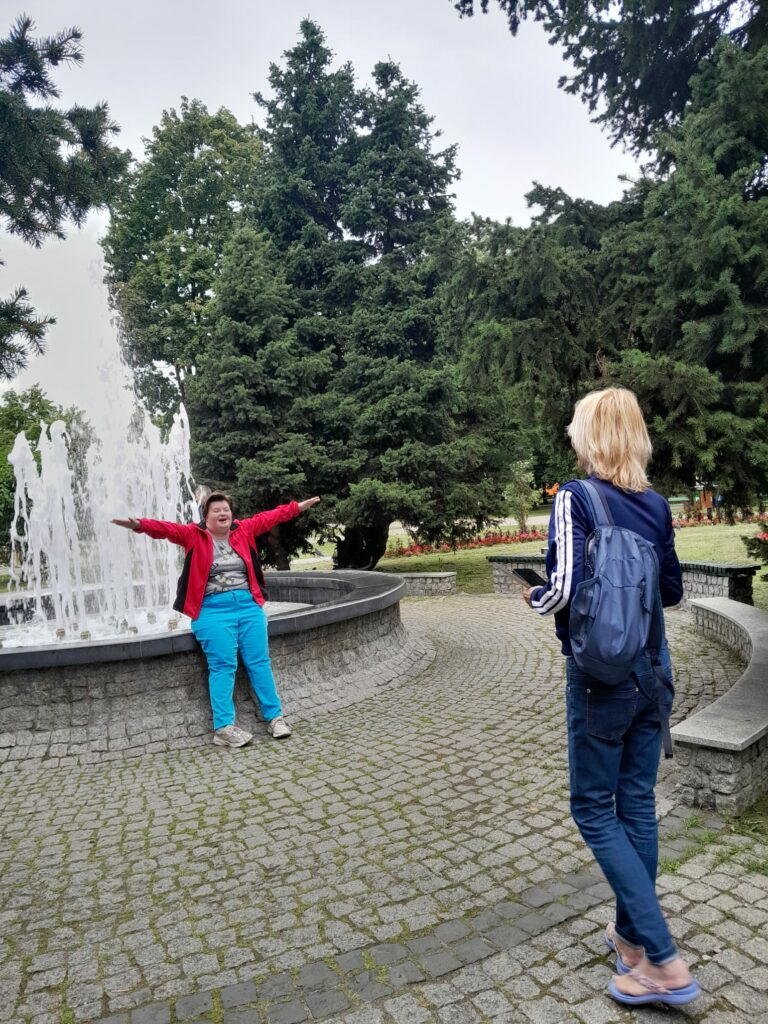 Osoba z niepełosprawnością pozuje do zdjęcia na tle fontanny. Naprzeciwko niej stoi kobieta.