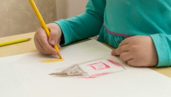 Ręce dziecka, które przy stoliku rysuje na kartce dom.