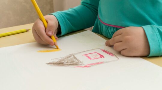 Ręce dziecka, które przy stoliku rysuje na kartce dom.