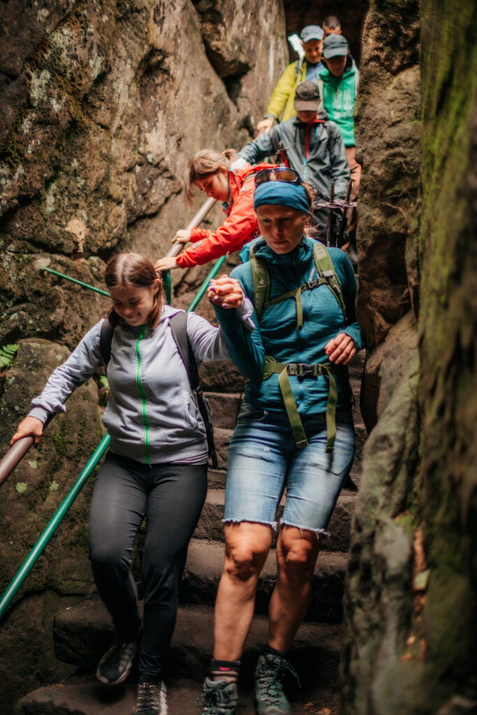Zdjęcie wykonane w górach, w czasie górskiej wyprawy. Grupa osób schodzi po kamiennych stopniach w skalnym korytarzu. Trzymają się poręczy. Wśród nich osoby z niepełnosprawnościami. Pracownicy fundacji Imago i wolontariusze (na pierwszym planie Agata Włodarczyk-Bucharowska) wpierają innych uczestników wyprawy). Agata trzyma mocno uczestniczkę za rękę, pomagając jej bezpiecznie zejść.
