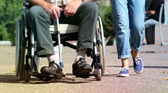 Zdjęcie wykonane na świeżym powietrzu. Widać na nim dwie osoby, które spacerują, jedna z nich jest na wózku dla osoby z niepełnosprawnością. Nie widać ich twarzy (zdjęcie od pasa/piersi w dół).