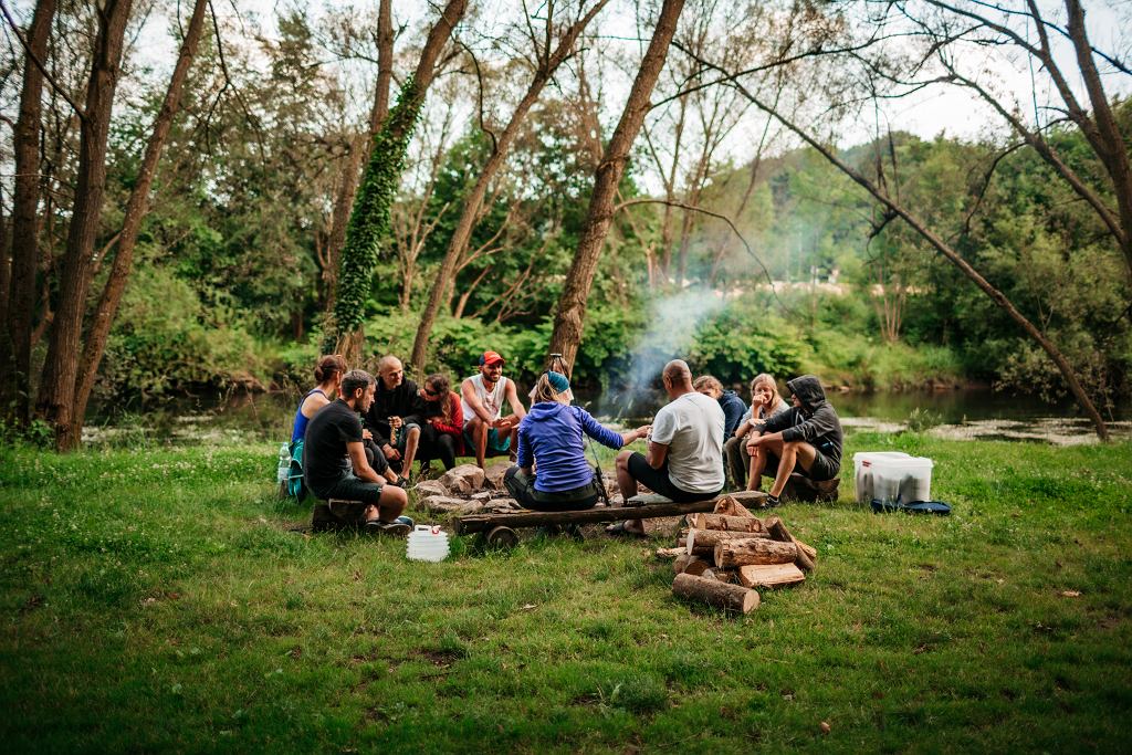 Polana w lesie, nad rzeką. Wokół ogniska siedzi grupa osób. Wokół jest zielono, zdjęcie mogło być wykonane wiosną lub wczesną jesienią.