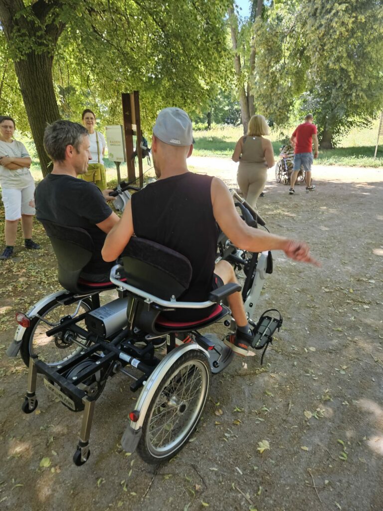 Dwie osoby siedzą obok siebie na rowerze typu tandem. Jedna z nich jest osobą z niepełnosprawnością ruchową. Zdjęcie wykonane w parku.